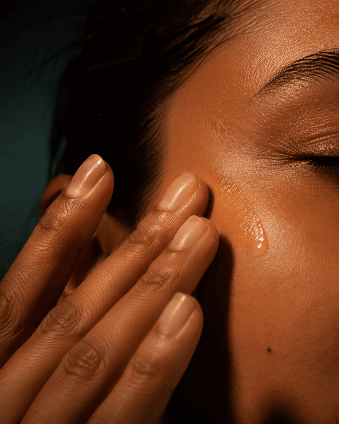 Fingertips pressing a clear facial oil into naturally textured skin in warm light